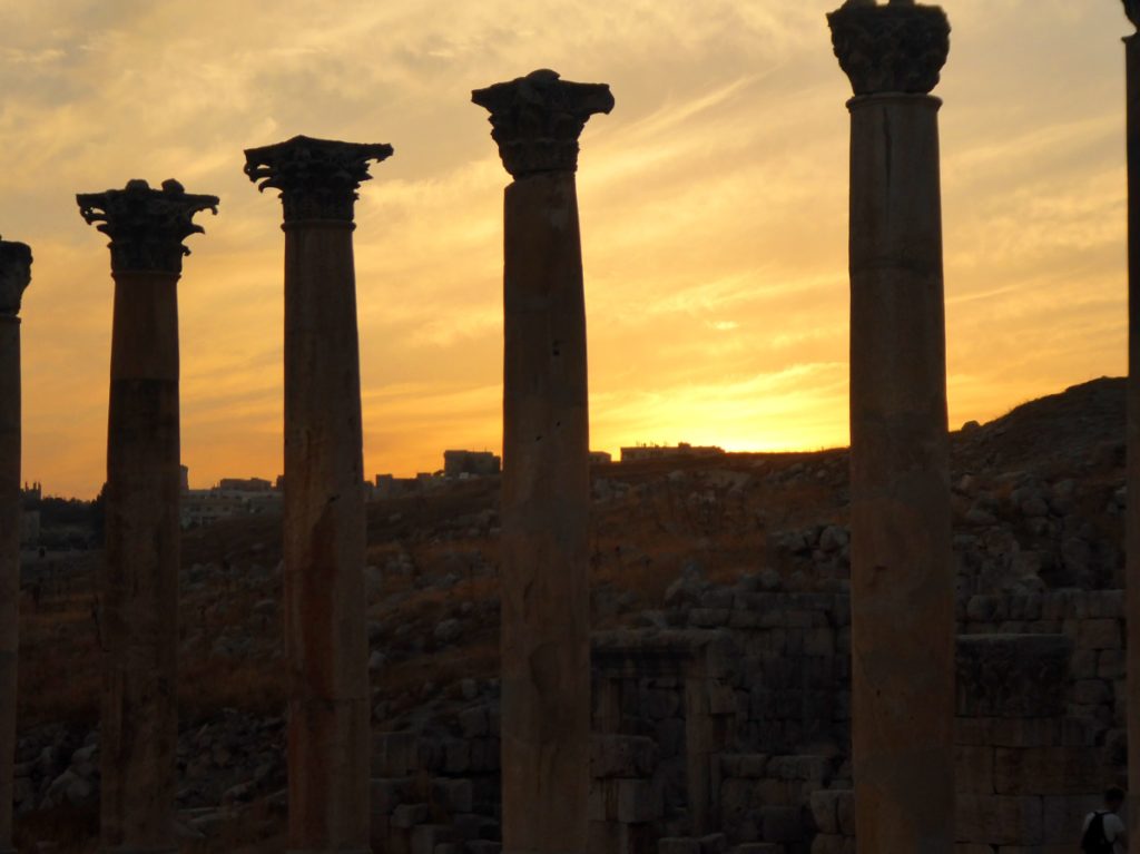 Die Sonne geht hinter vielen Säulen in Jerash unter, der Himmel hat ein paar dünne Wolken und ist gelb-orange