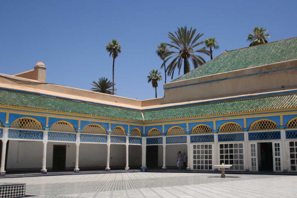 Eine der schönsten und wichtigsten Sehenswürdigkeiten in Marokko, der Bahia Palast in Marrakesch. Großer Innenhof mit hellen Bodenfliesen, umgeben von weißen Säulen, die von blauen Bögen überspannt werden. Grün gefliestes Dach. Kleiner Wasserbrunnen im Innenhof.