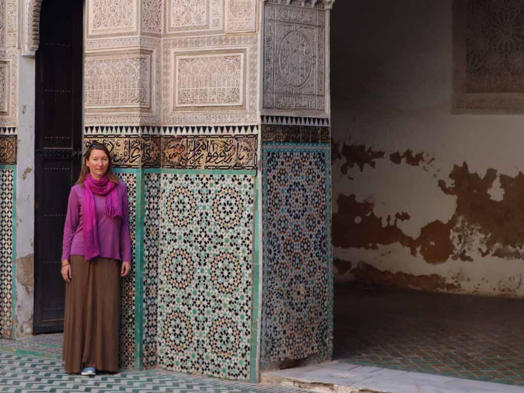 Women in long skirt and longsleeve shirt with pink scarf in front of tiled mosaic wall. What to wear in Fes, Morocco