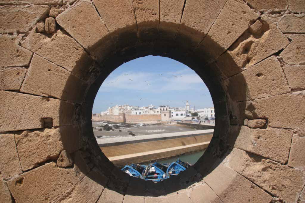 big round hole in a wall with view of fishing boats and a walled city. Essaouira, UNESCO sites in Morocco