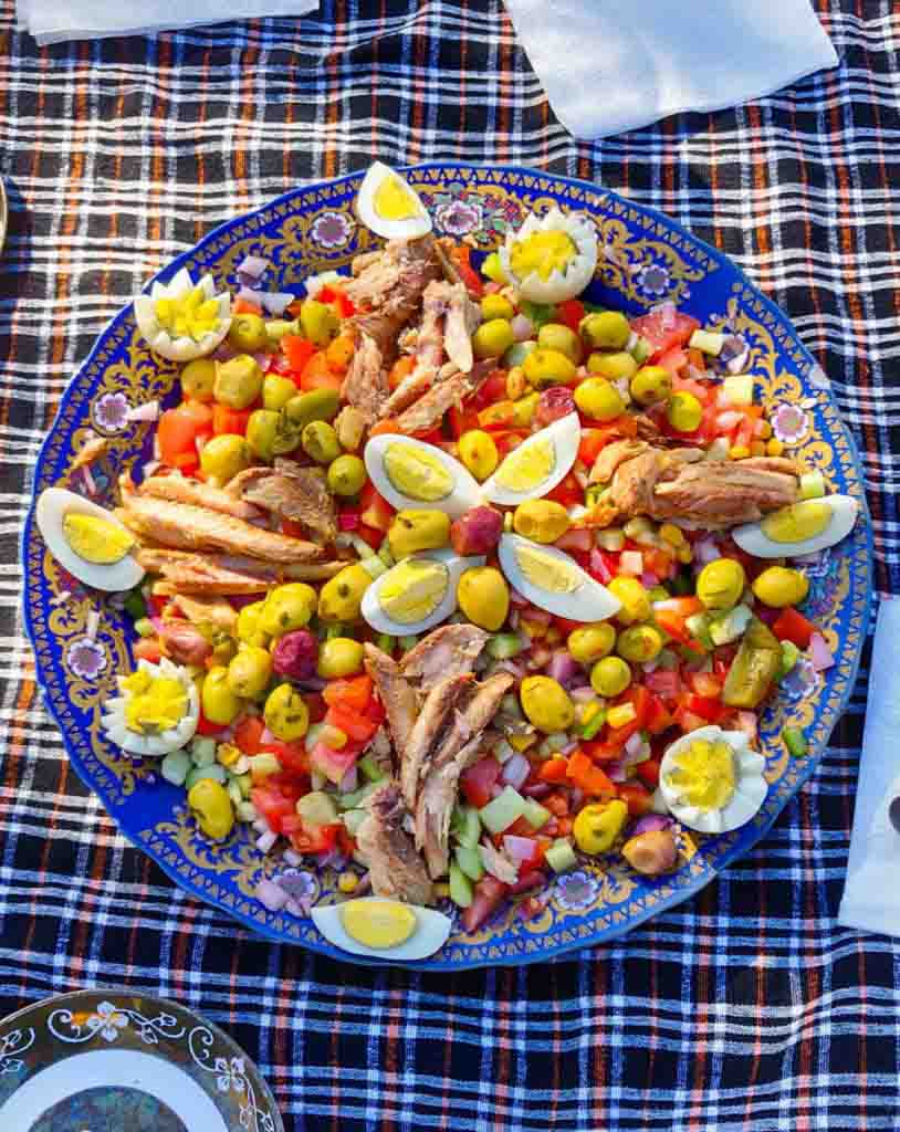 Typisch Marokkanisches Essen, ein marokkanischer Salat mit Tomaten, Gurke, Zwiebeln, Oliven, gekochten Eiern und Makrele aus der Dose. Alles auf großem Teller mit blau-goldenem Muster auf Picknickdecke.