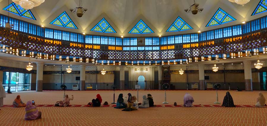 The main prayer hall in the Masjid Negara Malaysia, women sitting on the ground. blue and yellow windows on the upper levels