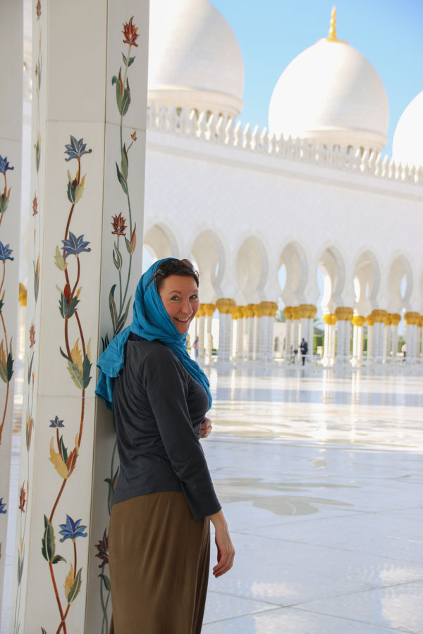 women in long skirt, long sleeve shirt and headscarf leaning on column and looking over her shoulder into camera. column decorated with mosaic flowers. Inside Sheikh Sayed mosque in Abu Dhabi, most beautiful mosque in the world