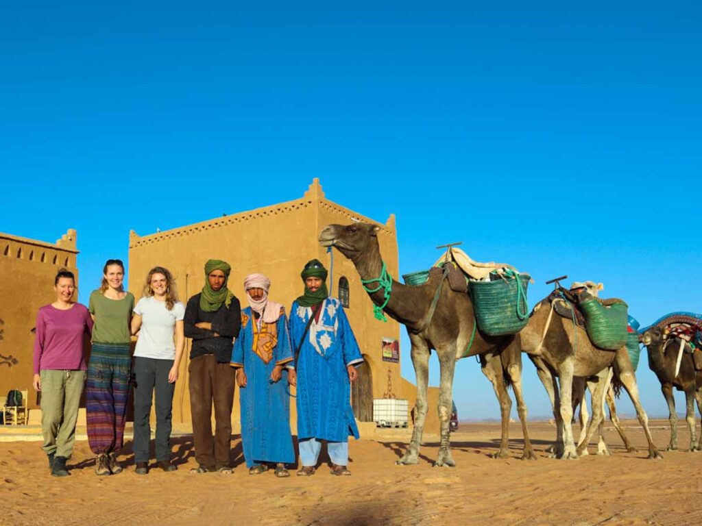 Group of 3 women and 3 men with 4 camels in front of typical moroccan clay building. Berber adventure tours, long distance desert hiking adventures in morocco 