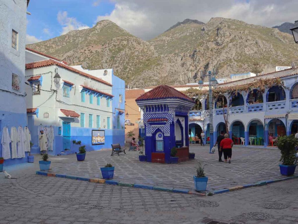 A water fountain with a red roof in the middle of a square. The fountain is tiled in blue and white mosaic. The houses painted blue and white. Blue pots with plants are places around in a circle. Mountains in the background. Things to do in Chefchaouen, Morocco