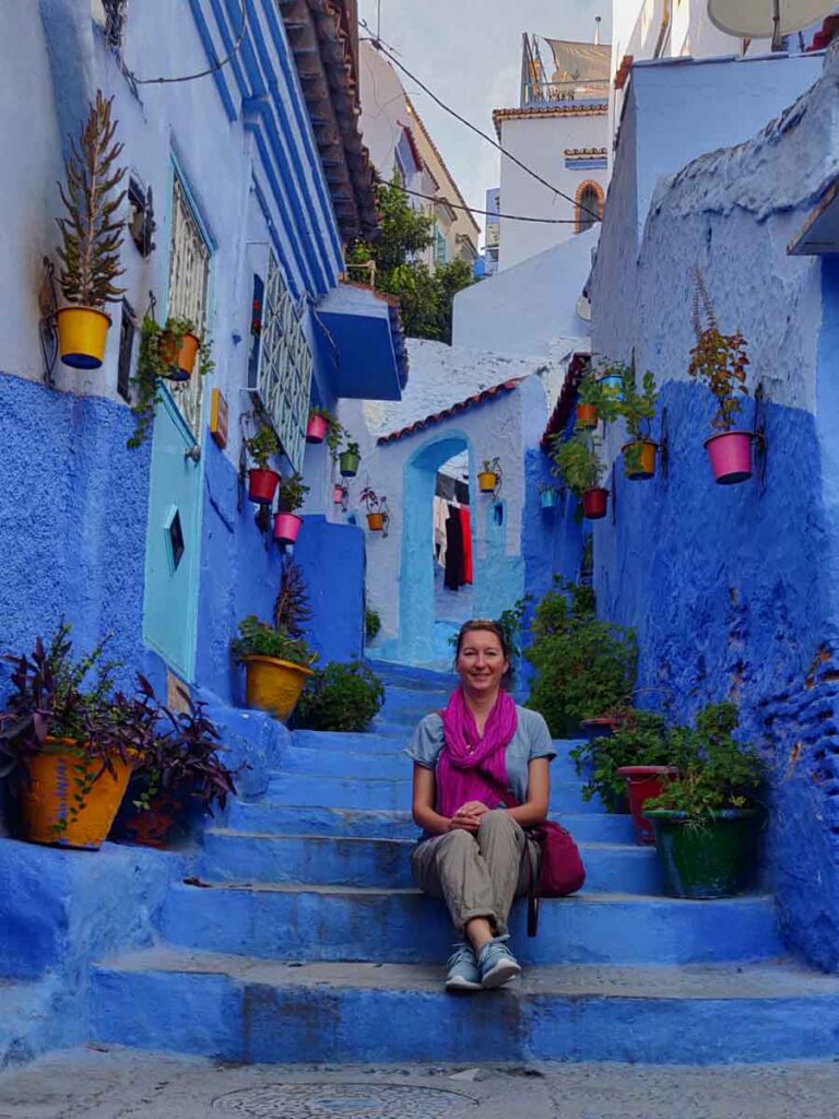 Smiling woman sitting on stairs leading upwards. Stairs and houses painted blue. Lots of plant and flower pots on the steps and walls. The woman is wearing a pink scarf, blue t-shirt and green pants. Instagram stairs of Chefchaouen, Morocco, things to do in Chefchaouen